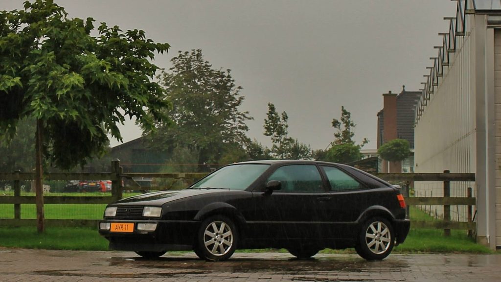 Afbeelding van een zwarte Volkswagen Corrado G60 in de regen met op de achtergrond een plattelandstafereel